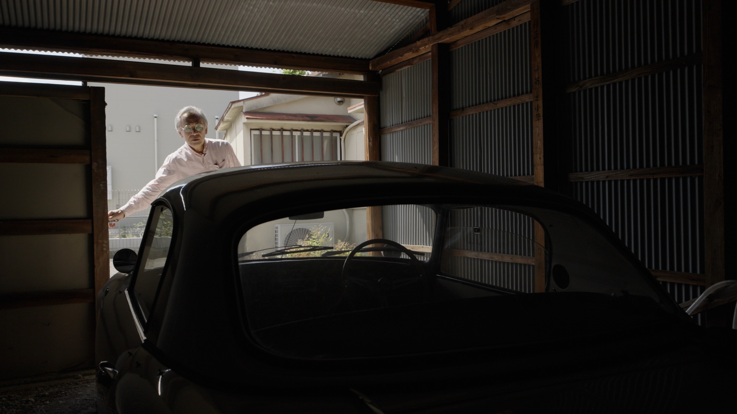 Tatsumi opening his garage door, a vintage car in shadow in the foreground