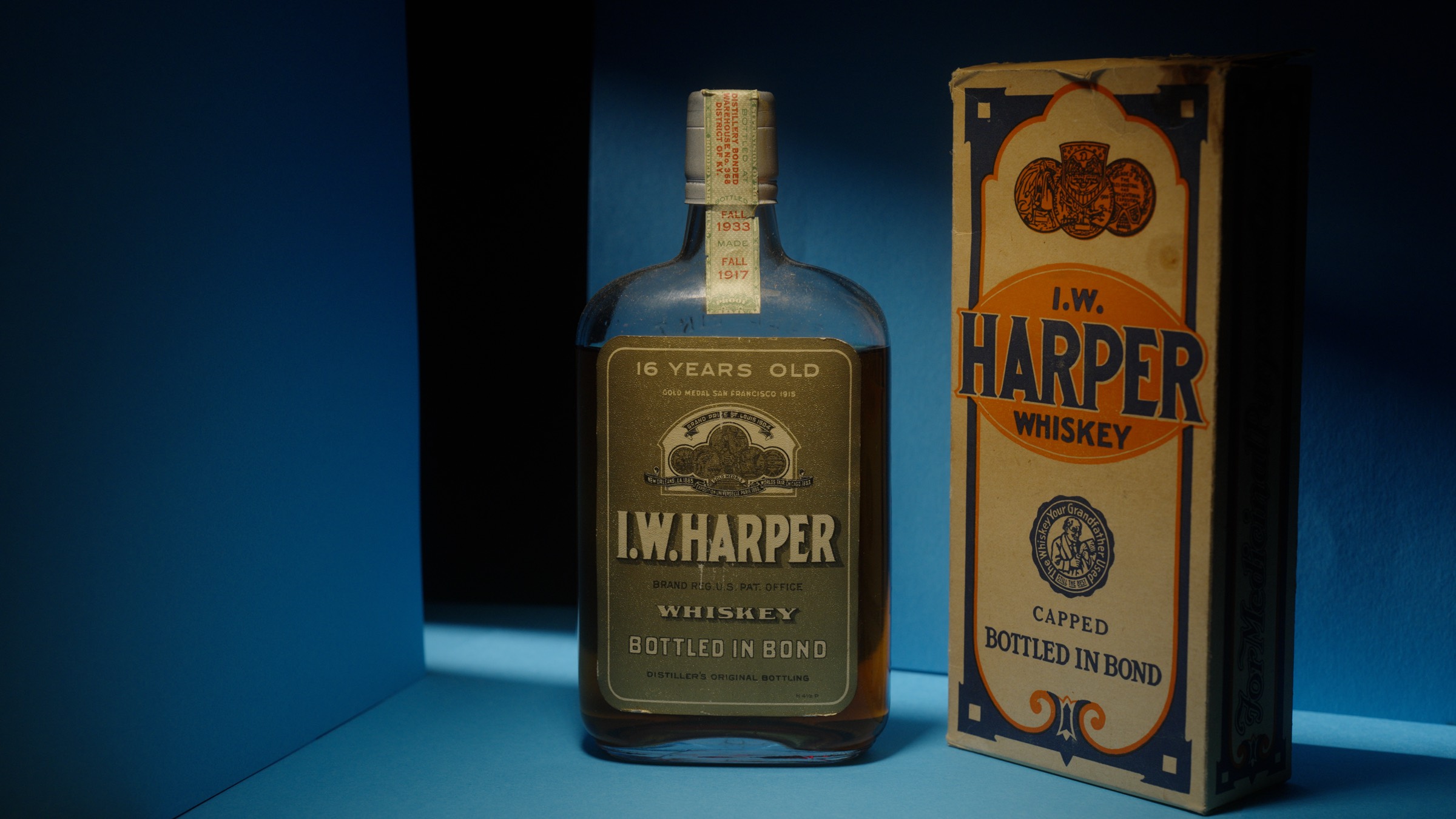 A bottle of I.W. Harper bottled-in-bond whiskey beside its original box, lit in deep blue