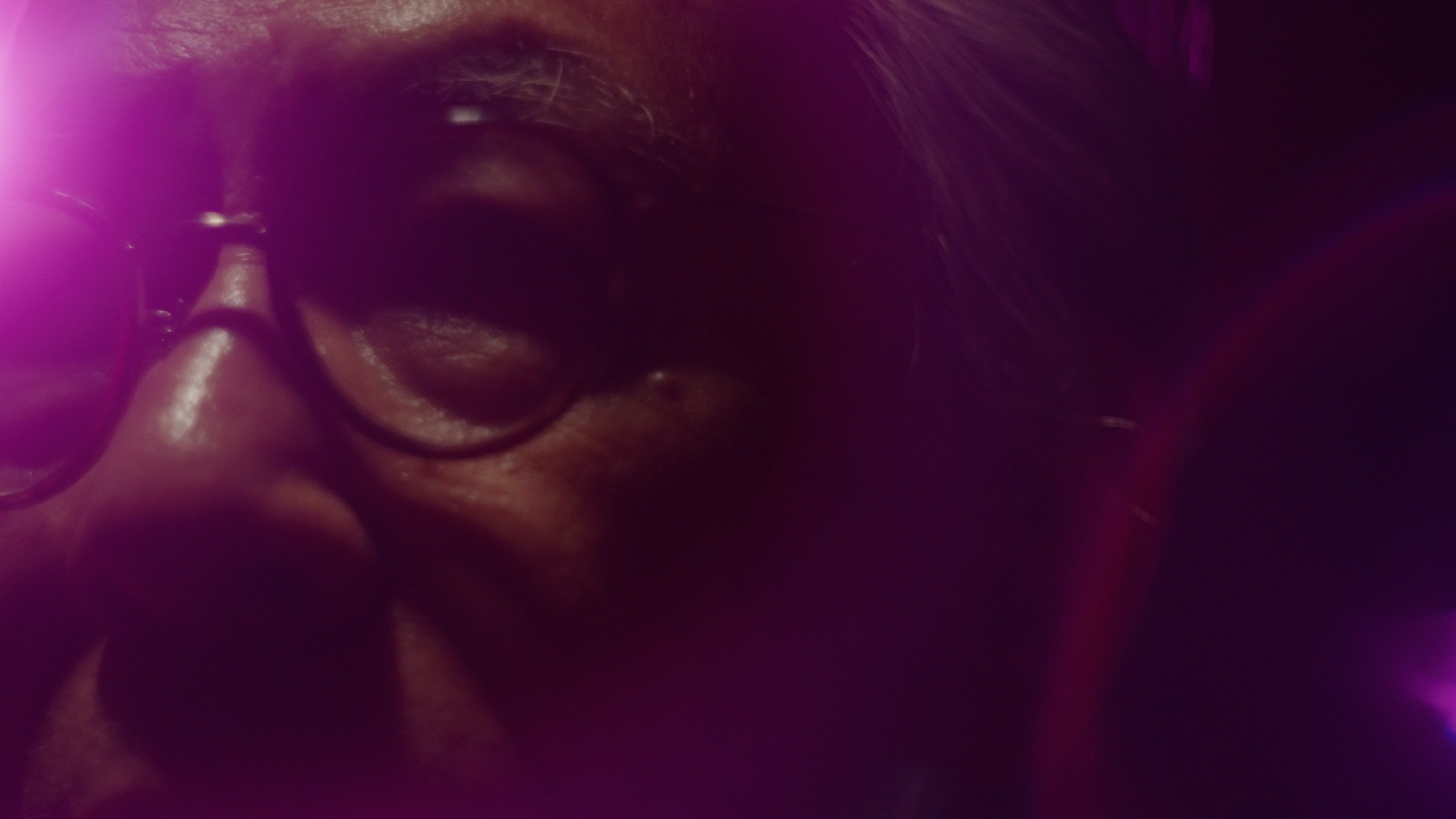 Seiichiro Tatsumi in close-up, his glasses and face washed in magenta neon