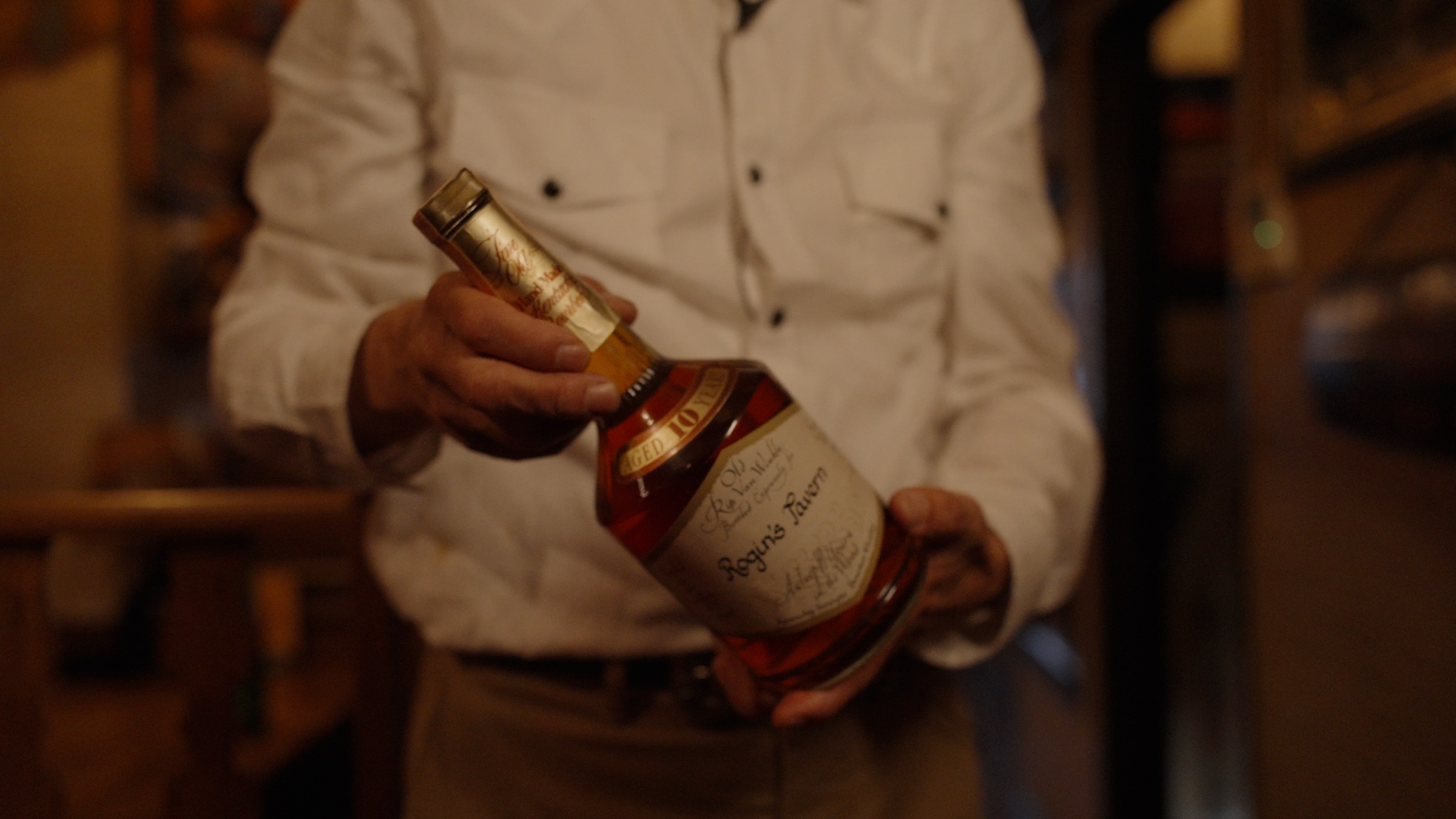 Hands holding a bottle labeled Rogin's Tavern in the warm light of the bar